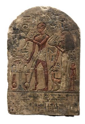 A round-topped limestone stela carved in raised relief, with traces of red, blue and black paint. On the left is an offering table piled with food, with tall jars beneath it. In the centre, Roma,  a b